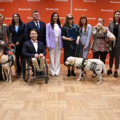 Grupa ludzi z psami asystującymi, jeden pies bez oka, w tym pies w wózku.