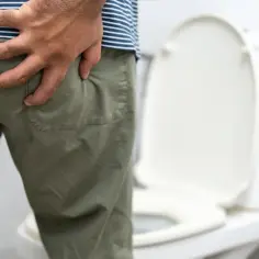 Mężczyzna trzyma się za pośladek, czując dyskomfort. Obok stoi toaleta, sugerując problemy z wypróżnianiem, być może związane z białymi kulkami w kupie.