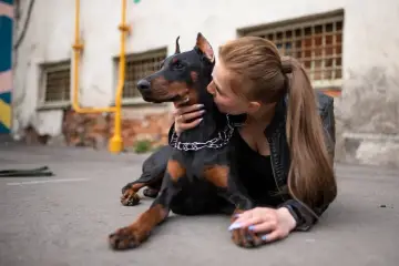 Doberman i młoda kobieta okazują sobie czułość. Pies leży na ziemi, a kobieta całuje go w policzek.