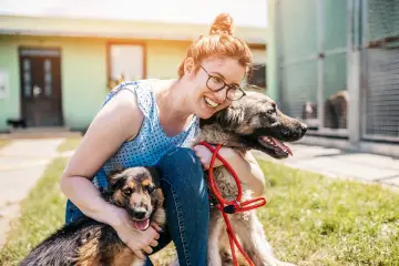 Uśmiechnięta wolontariuszka tuli dwa psy w schronisku. Szukają domów, to **setery do adopcji**.